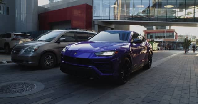 A sleek purple luxury sports car parked on a modern city street with other vehicles and contemporary buildings in the background.