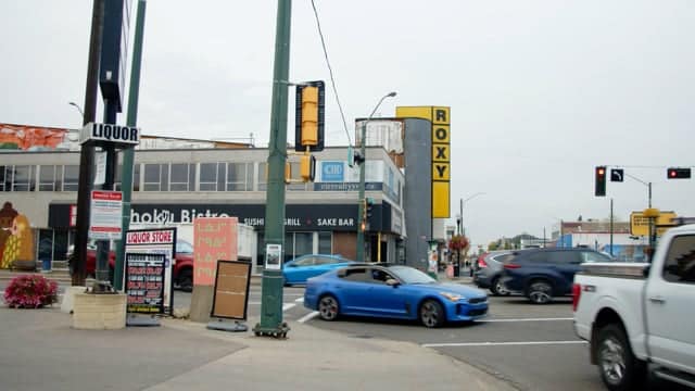 Blue sports car driving through busy city intersection with traffic lights and storefronts, showcasing urban nightlife and street scenes.