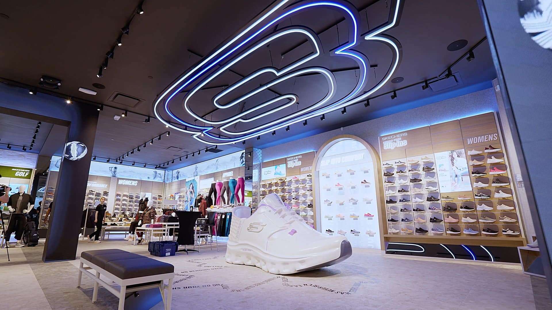 Brightly lit footwear store display with a giant white sneaker, shoe racks for men and women, and modern neon cloud ceiling design, showcasing trendy athletic shoes and sneakers.