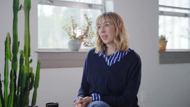 A woman with glasses and wavy blonde hair sitting indoors in a modern office or studio setting.