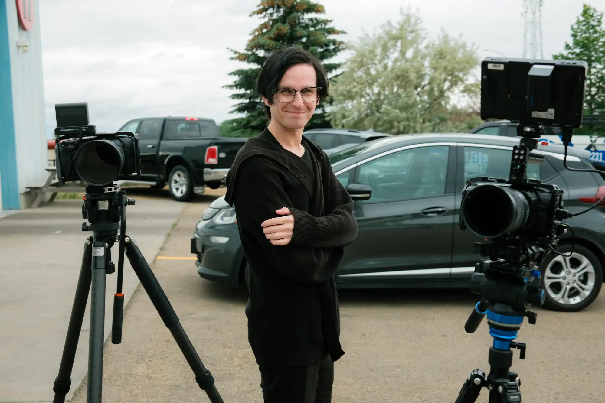 Glamorous young man standing outdoors with professional video cameras, ready for filming, in a parking lot with cars and trees, media production, filmmaking, videography, cinematography, Q Filmz Media relevance.