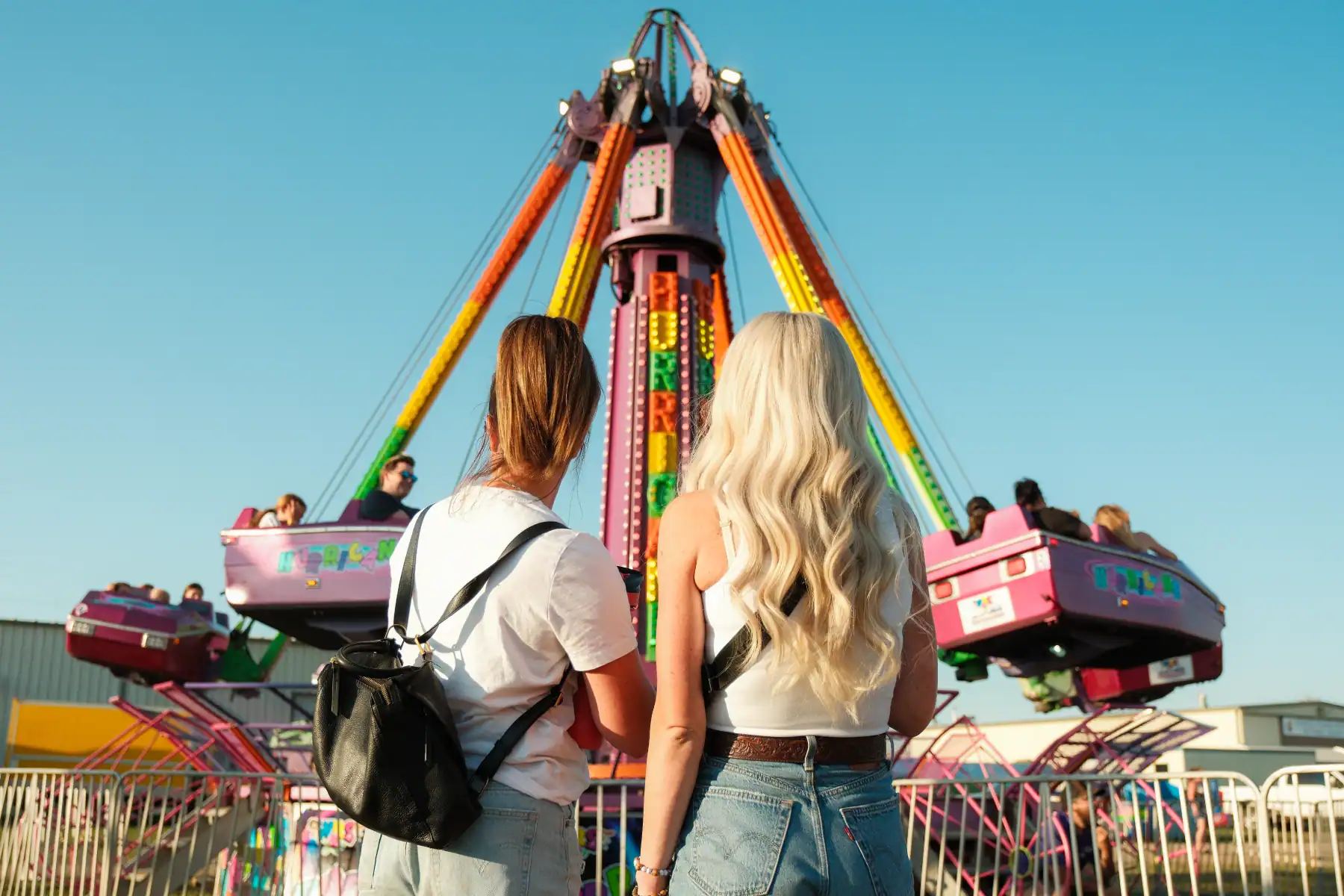Colorful amusement park ride spinning under a clear blue sky with two young women watching; vibrant carnival atmosphere, fun, entertainment, and leisure.