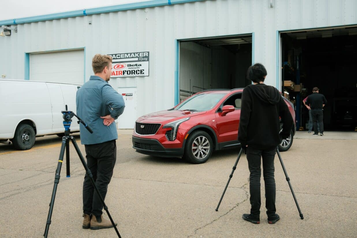 High-quality image of filmmakers filming a red Cadillac SUV outside an industrial building, showcasing modern automotive photography and video production for car commercials or promotional content.