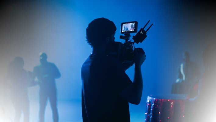 Filmmaker operating a professional camera on set with crew members in the background, illuminated by blue lighting.