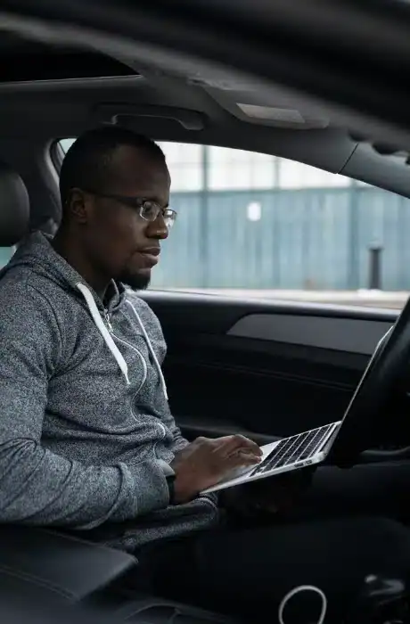 Young man with glasses using a laptop inside a modern car, focused on work or browsing.