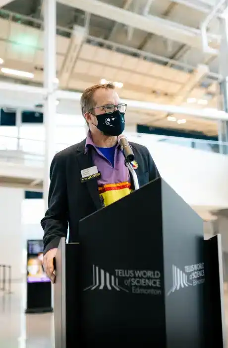 A man wearing glasses and a face mask speaking at a podium during an event at TELUS World of Science Edmonton.