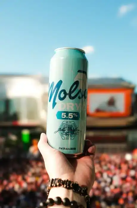A hand holding a Molek Dry 5.5% alcohol beverage can outdoors with a crowd and stage in the background.
