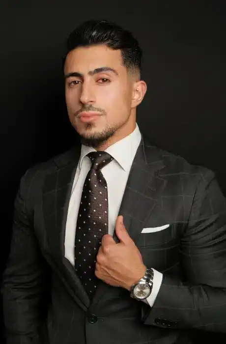 Confident man in a dark suit with a white shirt and polka dot tie, posing against a black background, showcasing style and professionalism.