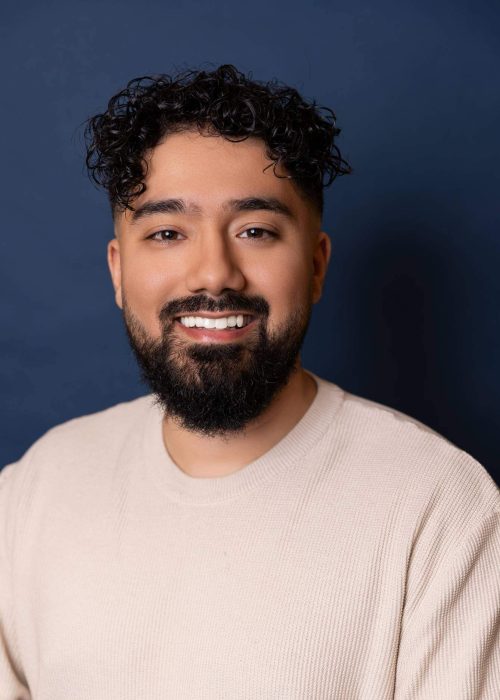 Smiling young man with curly hair and a beard against a dark blue background, showcasing confident and friendly personality, ideal for personal branding or professional photos.
